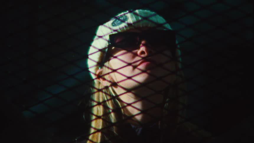 Close-up vintage shot of a stylish young woman in a beanie and sunglasses looking up, her face partially obscured by the shadow of a chain link fence