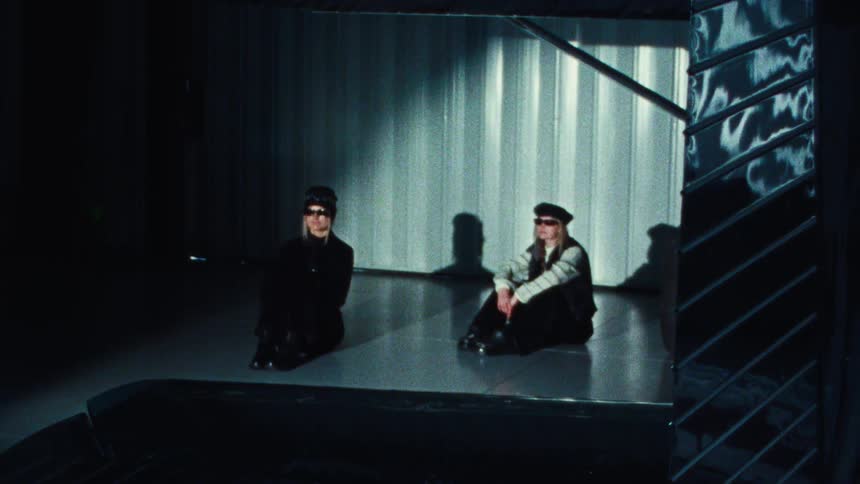 Super 8 film shot of two stylish blonde women in sunglasses and hats sitting on the floor of a dark, industrial room