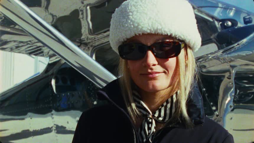 A stylish blonde woman in a fuzzy hat and sunglasses stands confidently in front of a reflective vintage airplane