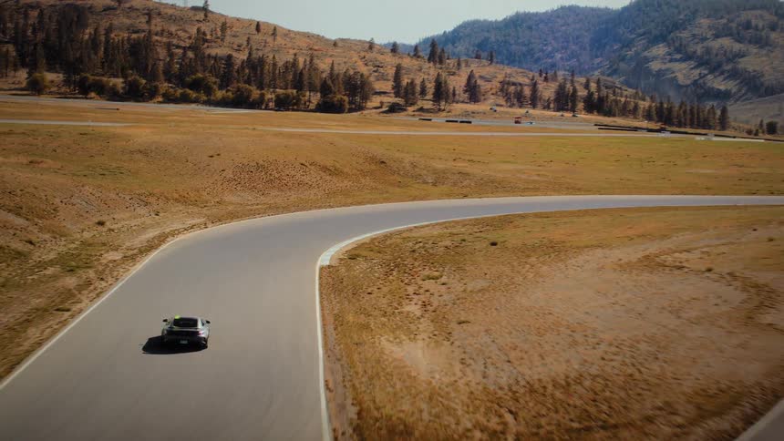 A fast aerial shot reveals a silver and green sports car speeding around a winding racetrack through a dry, hilly landscape