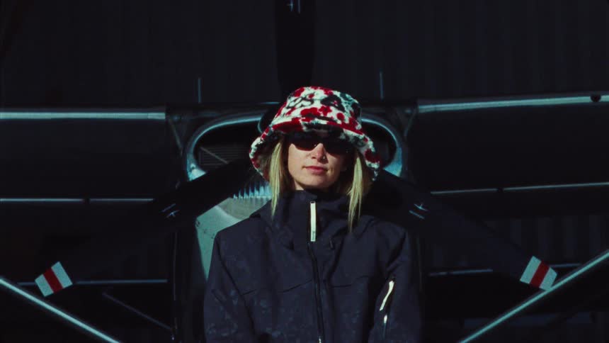 A stylish woman in a bucket hat and sunglasses confidently poses in front of a small propeller airplane