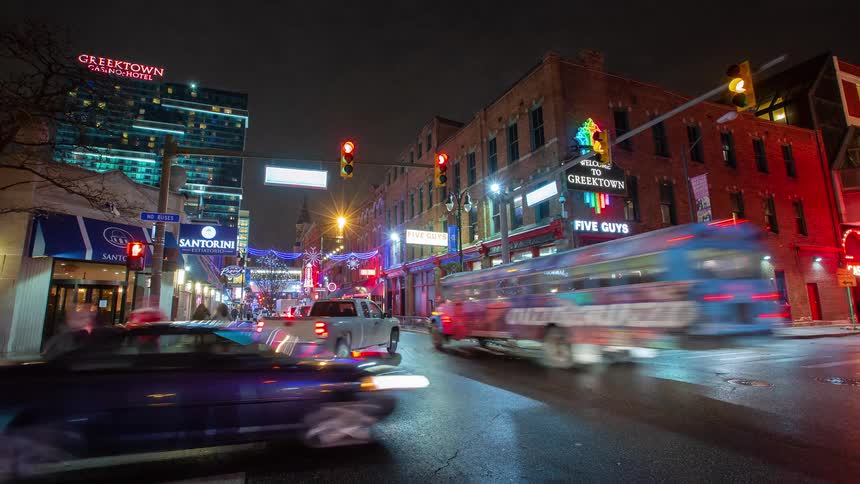 A vibrant time-lapse captures the bustling nightlife of Detroit's Greektown, with car light trails and pedestrians moving under colorful neon signs