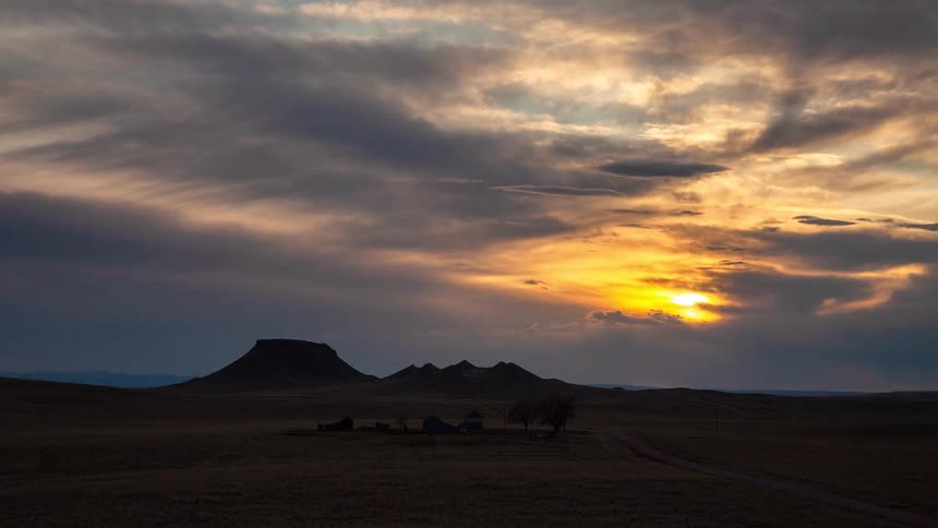 A dramatic timelapse shows clouds moving across the sky as the sun sets over a silhouetted, abandoned homestead and distant buttes on the open plains
