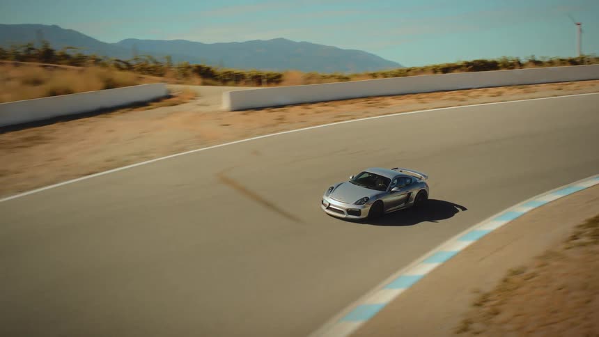 An exciting aerial tracking shot reveals a silver sports car speeding around a winding race track in a sunny, mountainous landscape