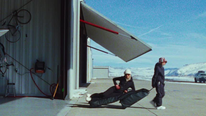 A woman in a snowsuit pulls her friend, who is playfully riding on a large wheeled snowboard bag, out of an airplane hangar on a sunny winter day