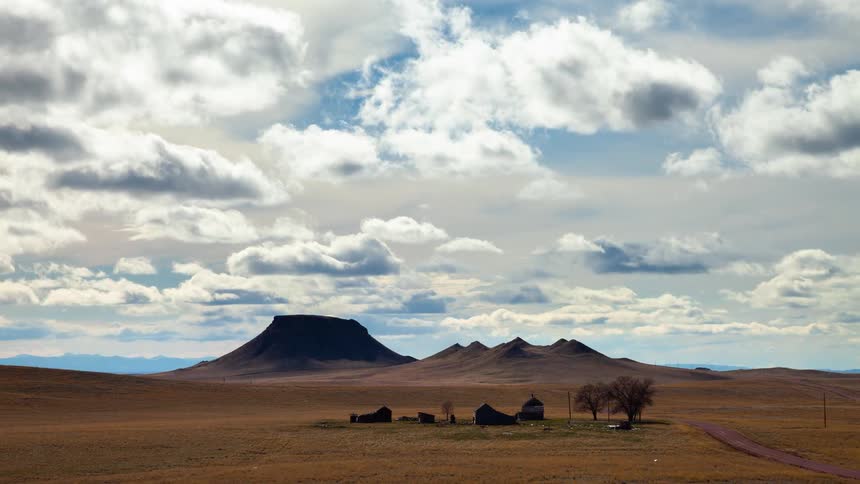Time-lapse of dramatic clouds and their shadows moving across a vast prairie landscape with an old homestead and a large butte in the distance