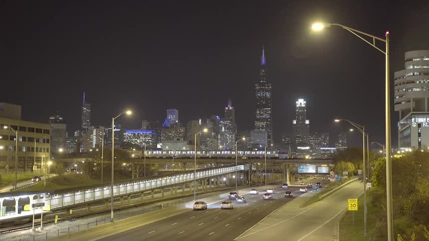 Cars and trains move along a busy expressway with the illuminated Chicago skyline in the background at night