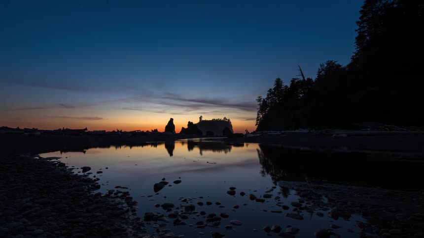 Time-lapse of a stunning sunset fading into a starry night sky over silhouetted sea stacks, reflected in the calm waters of a rocky beach