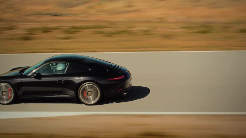 A sleek black sports car speeds along a winding racetrack through a dry, sun-drenched, hilly landscape