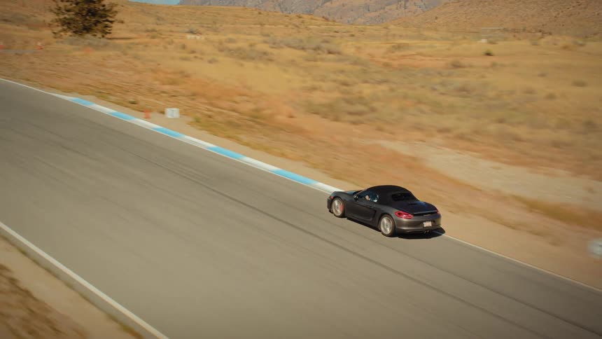 Aerial tracking shot of a grey convertible sports car driving at speed on a winding racetrack through a dry, sunlit, mountainous landscape