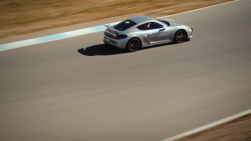 An aerial view tracks a silver sports car as it speeds around a professional racetrack in a rural setting