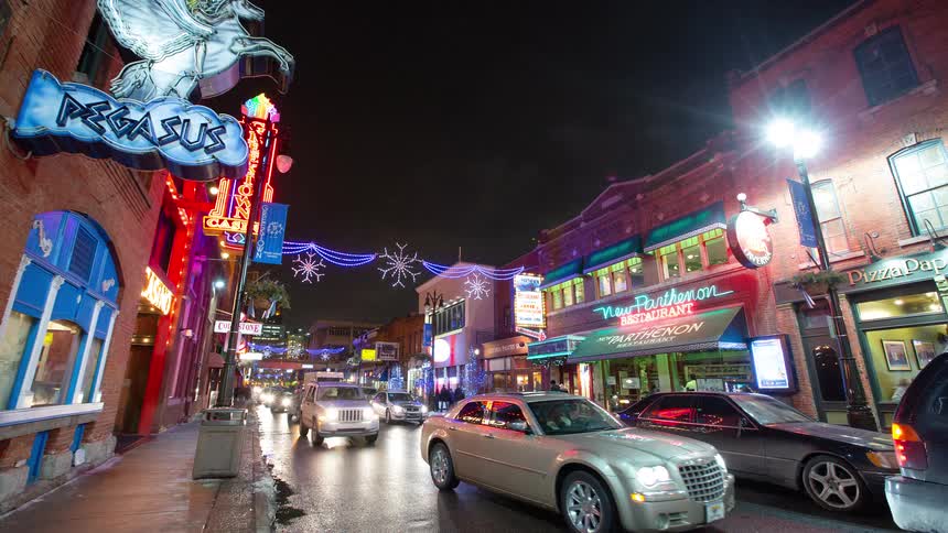A nighttime timelapse of a bustling city street in Detroit's Greektown, with neon signs, holiday lights, and heavy car and pedestrian traffic