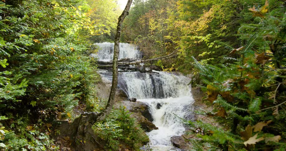 A beautiful multi-tiered waterfall cascades through a lush autumn forest as a gentle rain shower begins, catching the sunlight