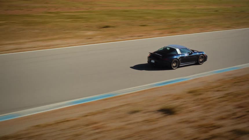 A sleek black sports car speeds along a professional racetrack surrounded by a dry, hilly landscape on a sunny day