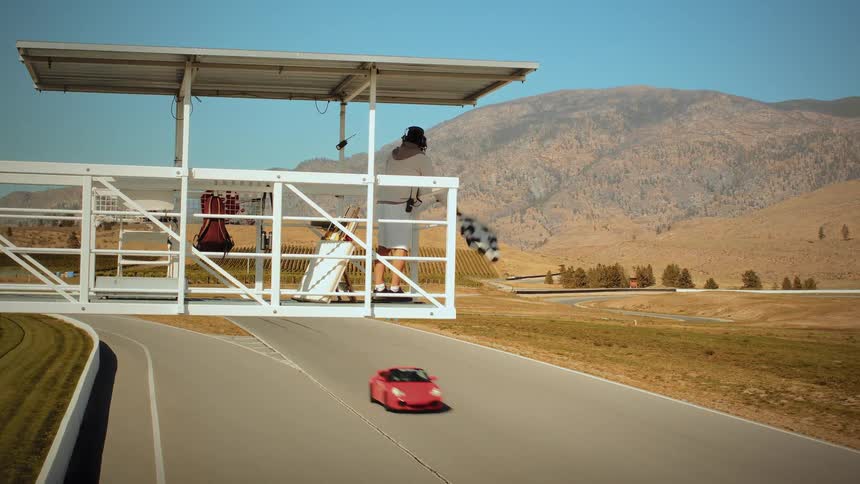 A man waves a checkered flag from an elevated platform as sports cars race below on a track set in a dry, mountainous landscape