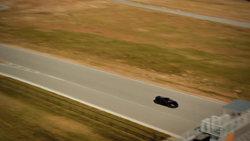 Aerial view of a sleek black sports car driving at high speed along a straightaway on a racetrack set in a hilly, rural landscape
