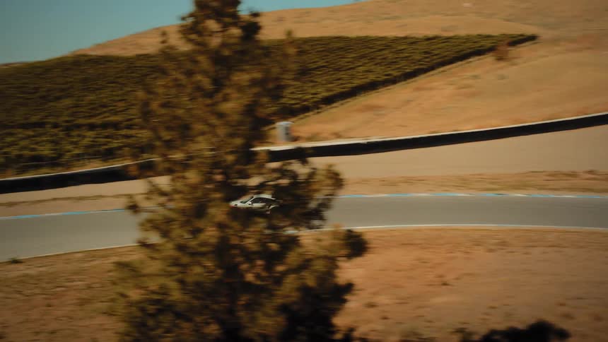 An aerial tracking shot follows a white Porsche as it races along a winding circuit through an arid, mountainous terrain