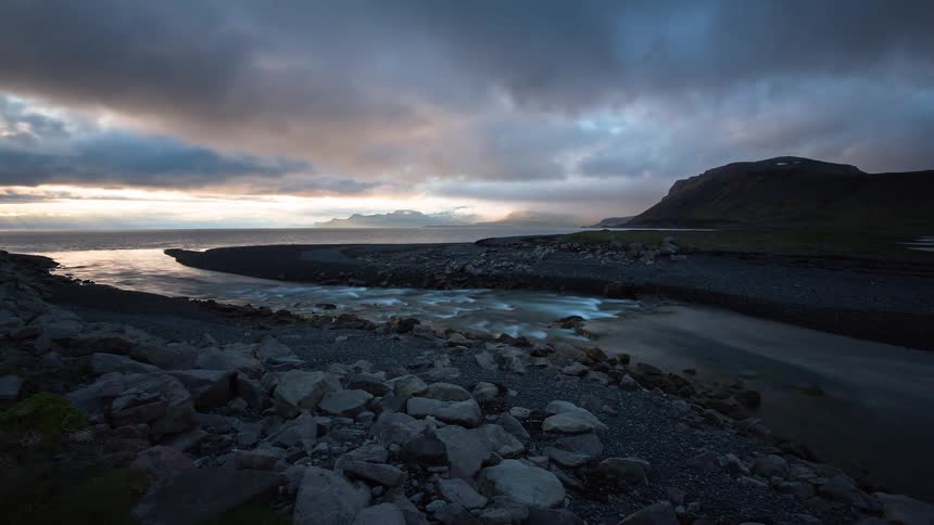 A dramatic time-lapse shows a river flowing into the ocean on a rocky coastline as clouds move across the sky at twilight