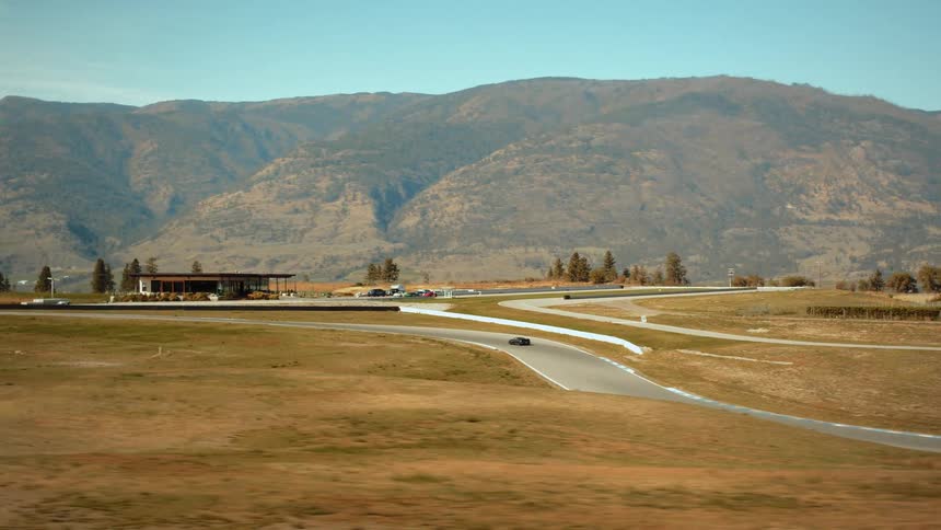 A wide tracking shot of a single race car driving on a racetrack set in a scenic mountain valley on a sunny day