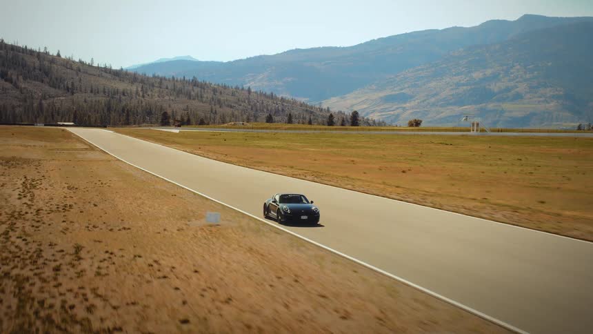 An aerial view follows a sleek black sports car as it gracefully navigates a turn on a racetrack set against a backdrop of rolling hills and mountains
