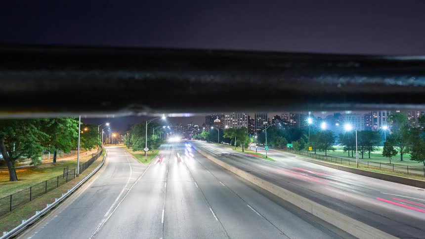 A smooth motion-controlled time-lapse reveals a bustling highway at night with the city skyline in the background, starting from behind a flower box on a bridge