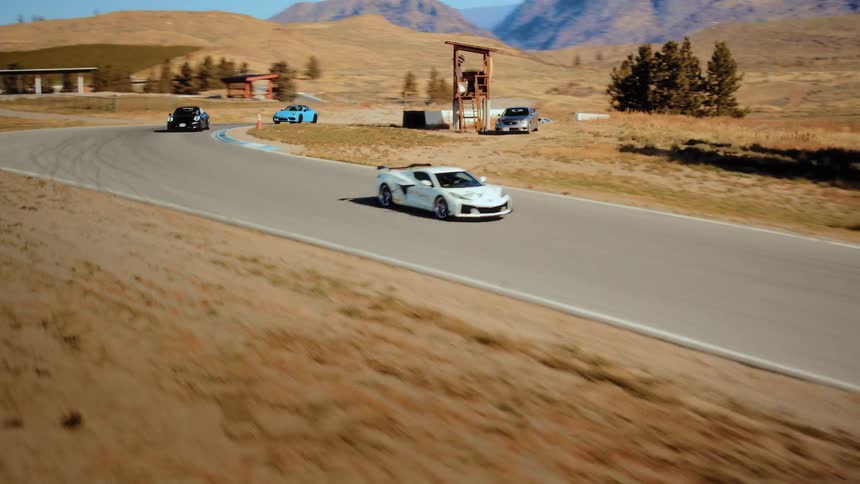 Aerial view of a white Chevrolet Corvette leading other sports cars around a winding race track in a sunny, arid mountain landscape