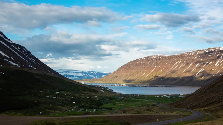 A scenic timelapse of clouds moving over a small town nestled in a valley by a fjord between two majestic, snow-capped mountains in Iceland