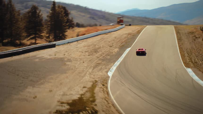 An aerial shot follows a sleek red sports car as it speeds around a curve on a racetrack in a scenic, arid mountain landscape