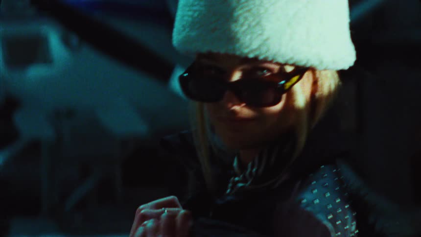 A stylish blonde woman in a white fuzzy hat and sunglasses smiles while looking over her shoulder in a dark, moody setting