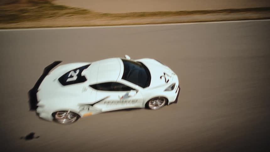 A thrilling FPV drone chase follows a white Corvette race car speeding around a winding track in the mountains