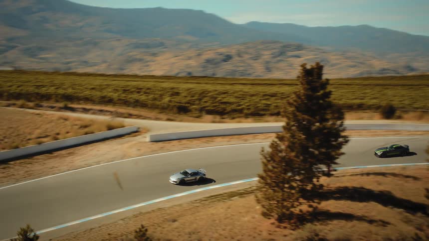 An aerial view of a sleek silver sports car as it speeds around a race track set in a dry, mountainous landscape