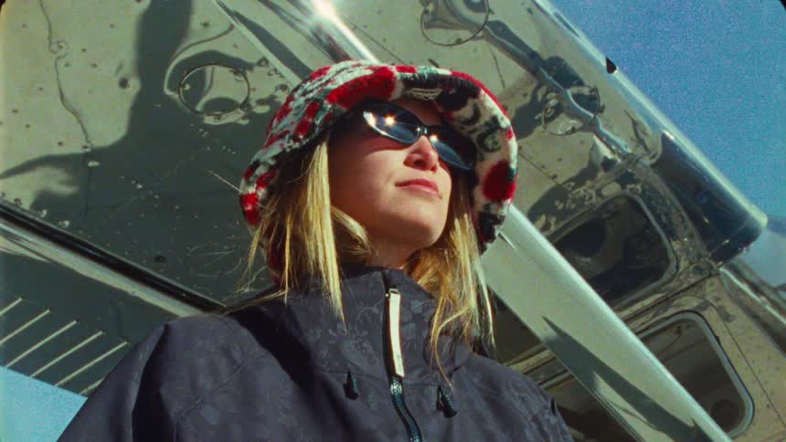 A confident woman in a bucket hat and sunglasses poses next to a shiny, silver airplane on a sunny day