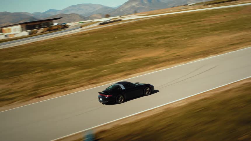 An aerial shot follows a sleek black sports car as it speeds around a winding racetrack set in a scenic, arid mountain landscape
