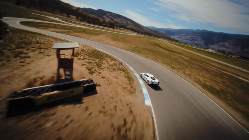 Dynamic aerial footage follows a white Corvette race car speeding around a winding track in a sunny, mountainous landscape
