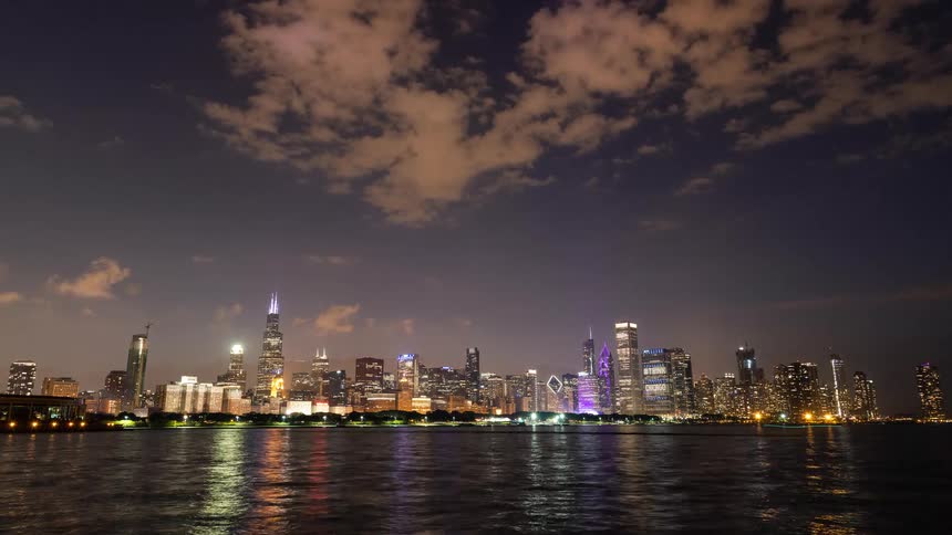 The Chicago city skyline twinkles at dusk as lights reflect on the calm waters of Lake Michigan and clouds drift by in a time-lapse