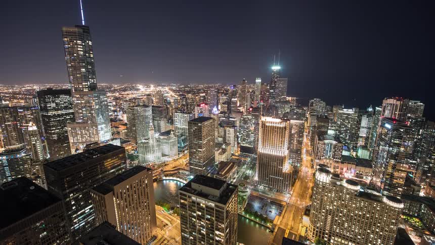 An expansive aerial time-lapse captures the vibrant energy of downtown Chicago at night, with light trails from cars and boats crisscrossing the cityscape