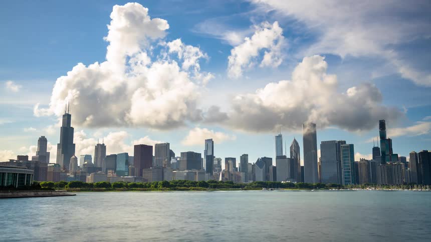 A bright daytime timelapse captures the iconic Chicago skyline as clouds drift over the skyscrapers and boats sail across Lake Michigan