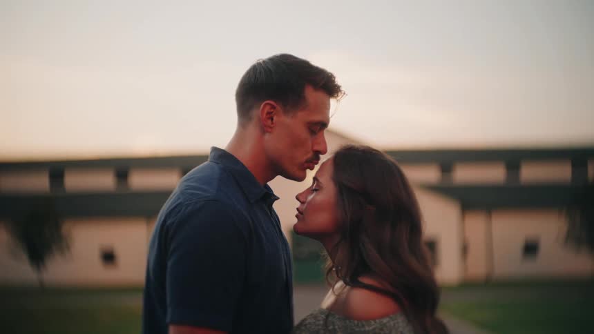 A man with a mustache gently kisses his girlfriend on the forehead in a tender moment at sunset in front of a rural stable