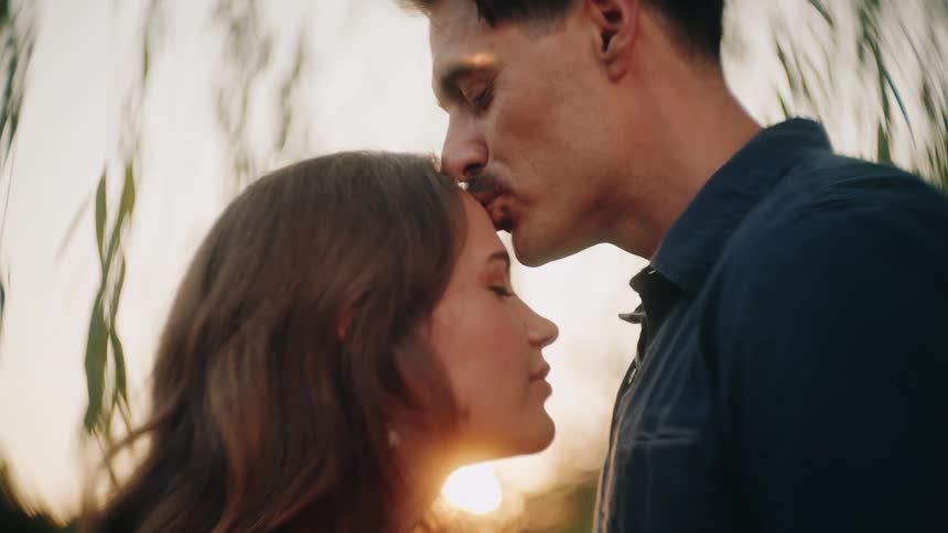 A man tenderly kisses a woman on the forehead in a slow-motion, romantic close-up during a beautiful sunset