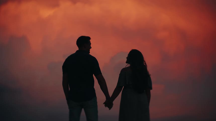 A romantic couple holds hands while silhouetted against a dramatic and fiery red sky at sunset