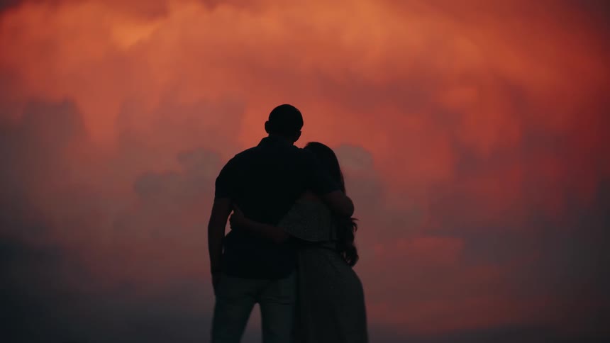 A silhouetted couple romantically embraces while watching a fiery orange sunset together in slow motion