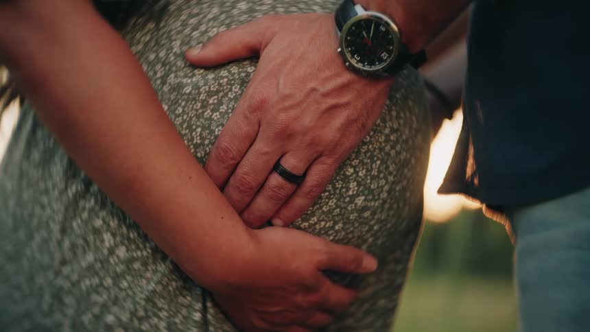 A close-up of an expectant couple's hands gently resting on the pregnant belly during a beautiful sunset
