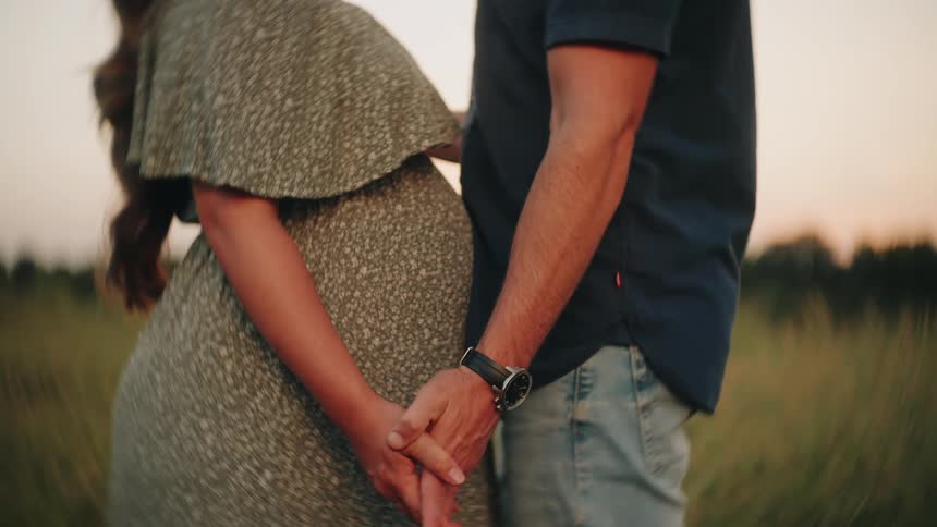 A loving couple expecting a baby tenderly hold hands while standing together in a field during a warm sunset