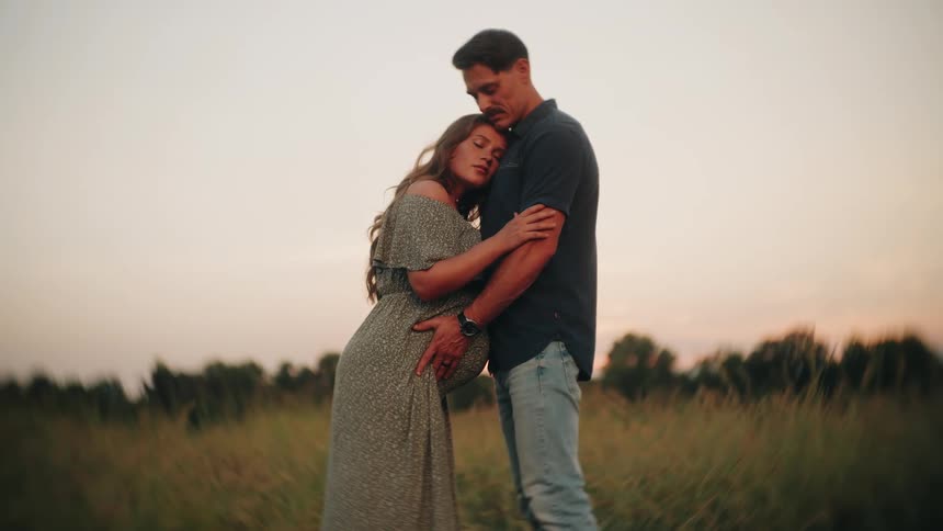 A pregnant woman rests her head on her partner's chest as they stand together in a beautiful golden field during sunset