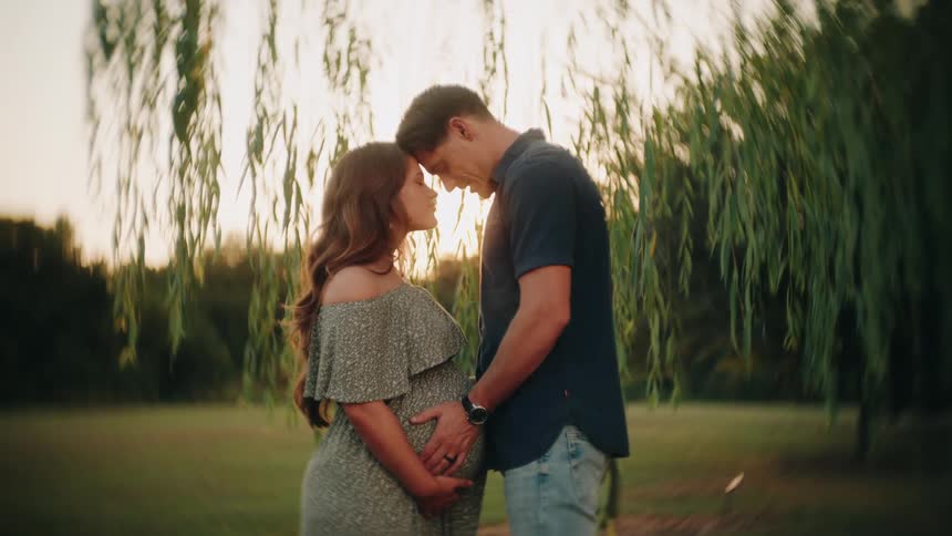 A pregnant woman and her partner share an intimate moment, foreheads touching, under a weeping willow tree as the sun sets