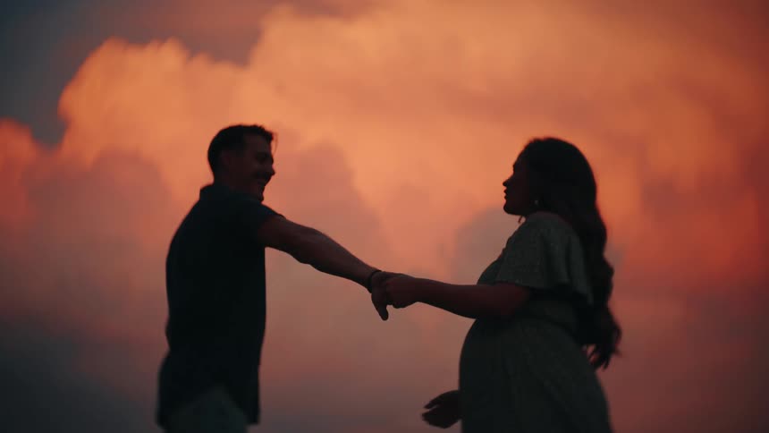 A silhouetted man and pregnant woman hold hands and dance slowly against a stunning and colorful sunset sky