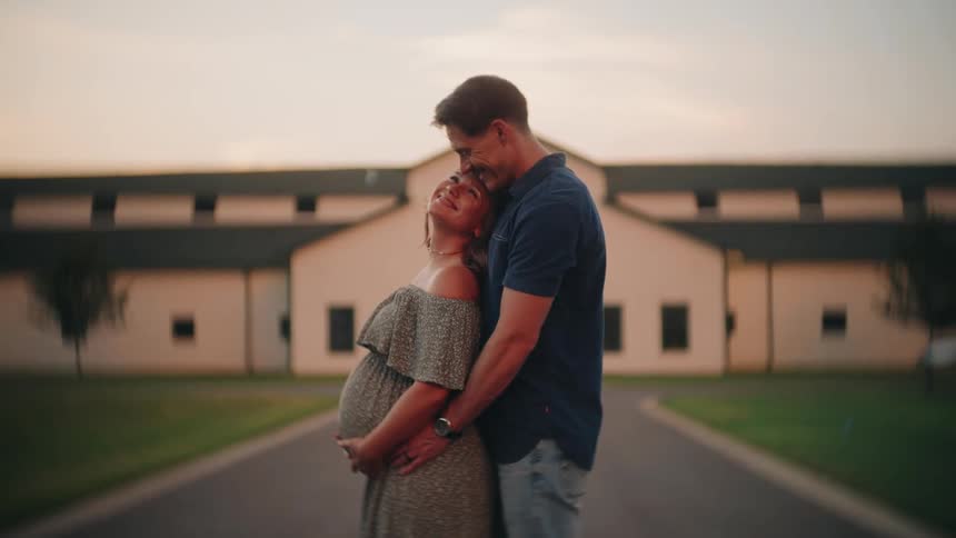 An expecting couple shares a loving embrace at sunset, with the man gently holding his pregnant wife's belly in front of a rustic farmhouse