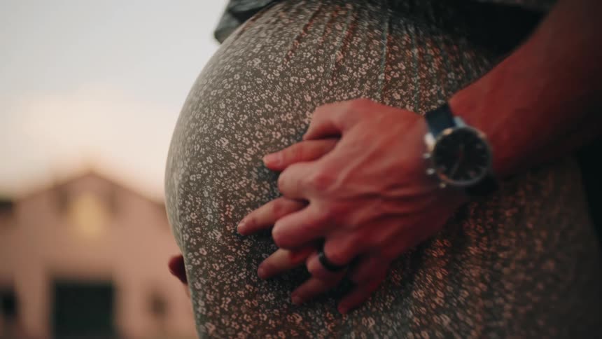 An intimate close-up shows a husband's hand over his wife's on her pregnant belly during a warm sunset
