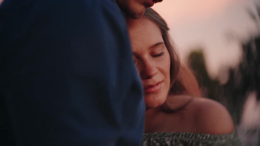 A young woman with her eyes closed peacefully rests her head on her partner's chest in a loving embrace at sunset