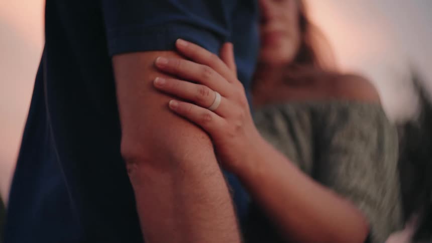 A woman's hand with a wedding ring gently caresses her partner's arm in a slow-motion, intimate moment at sunset